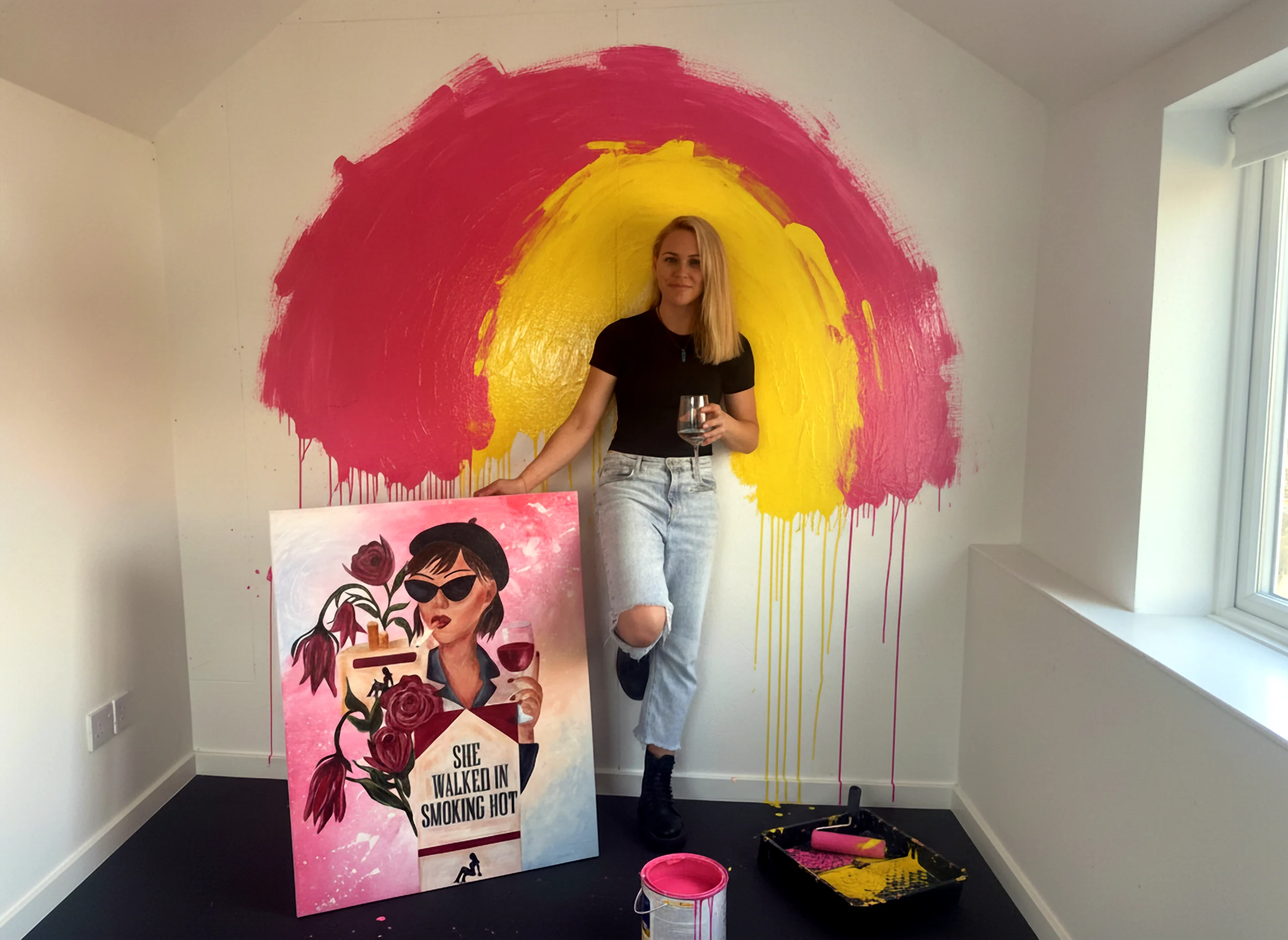 Rach Hesketh in her studio, the wall behind her rolled with pink and yellow paint, holding her painting She Walked In Smoking Hot
